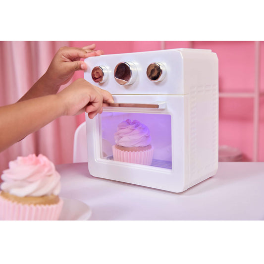A white toy oven with a transparent door showing a pink cupcake inside, with two hands turning dials on top. A play cupcake sits in the foreground on a pink table against a pink background.