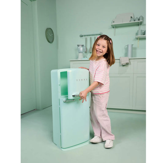 A young girl smiling and posing next to a retro-style mint green refrigerator in a matching pastel kitchen setting.