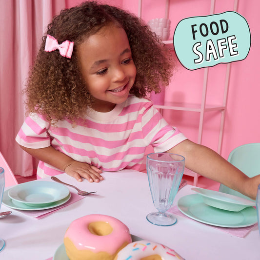 A young girl with curly hair wearing a pink and white striped shirt sits at a table with mint green dishes and donuts in a pink room. A light blue badge in the top right reads 'FOOD SAFE'.