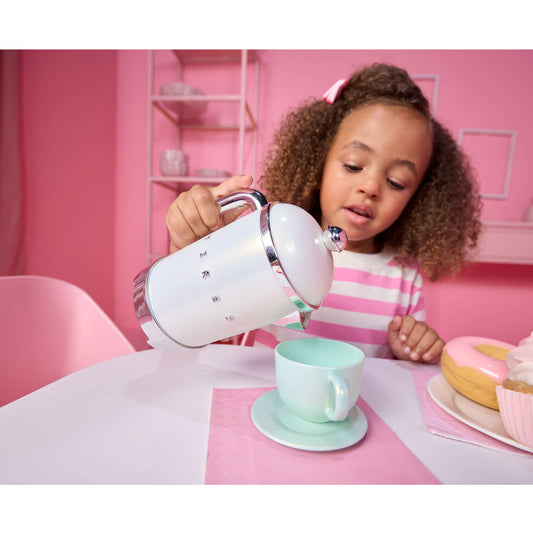 A young girl with curly hair pours water from a white kettle with chrome accents into a pastel mint green teacup on a saucer, set in a pink-themed room with a pink striped shirt and a pink frosted donut on the table.