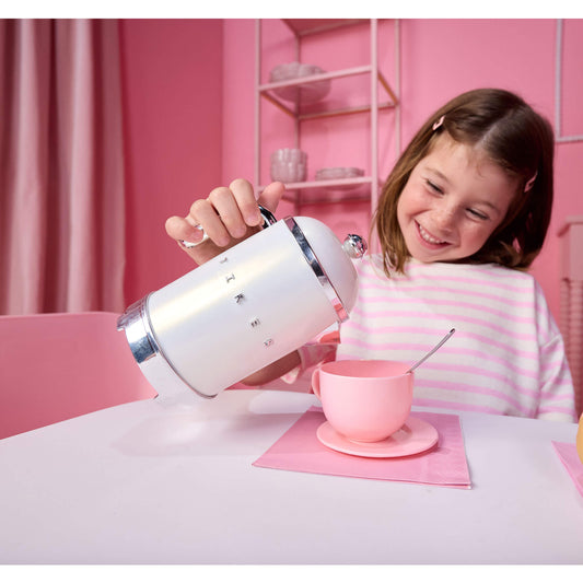 A young girl smiling while pouring water from a white and chrome kettle into a pink teacup and saucer on a table, set against a pink room background.