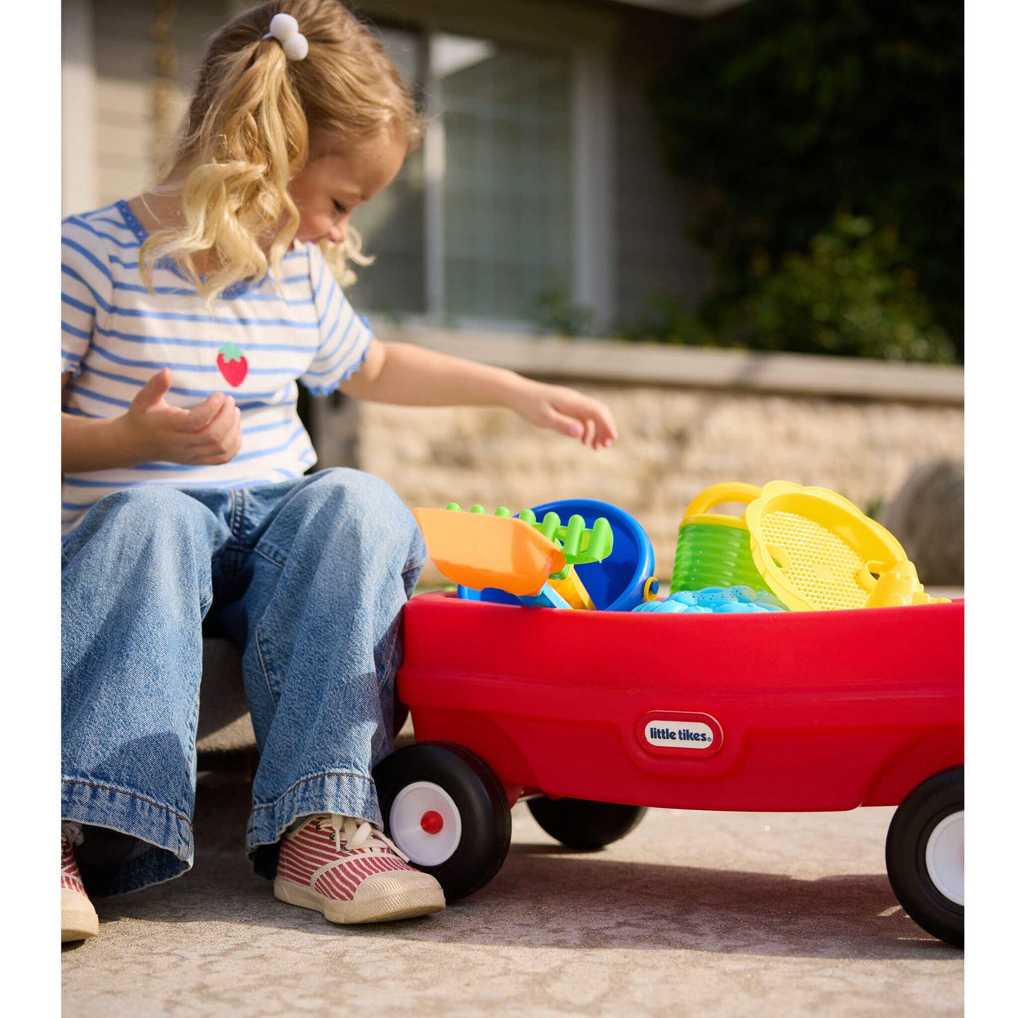 Child playing with a red toy wagon filled with colorful sand toys outdoors.