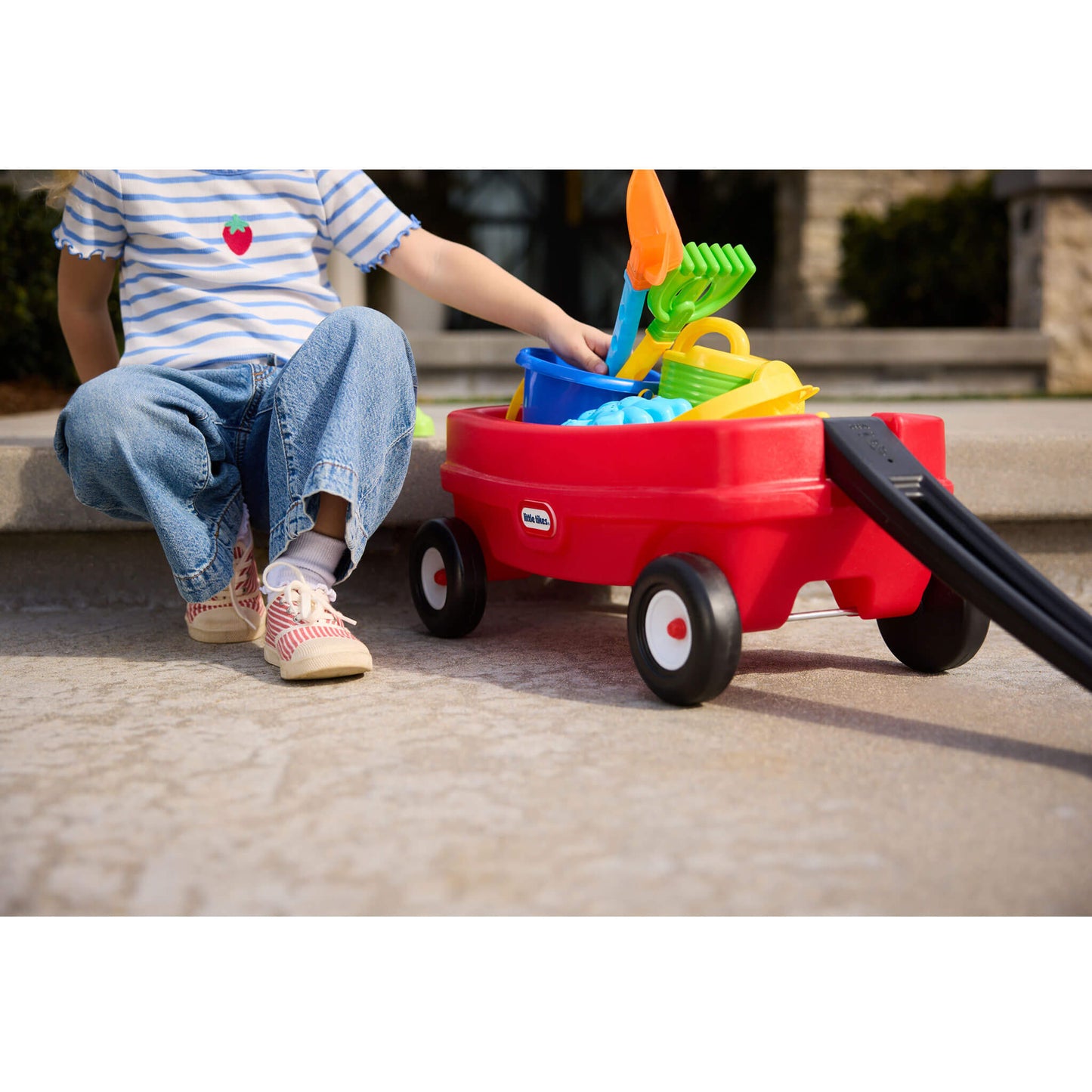 Child playing with a red toy wagon filled with sand toys on a concrete surface.