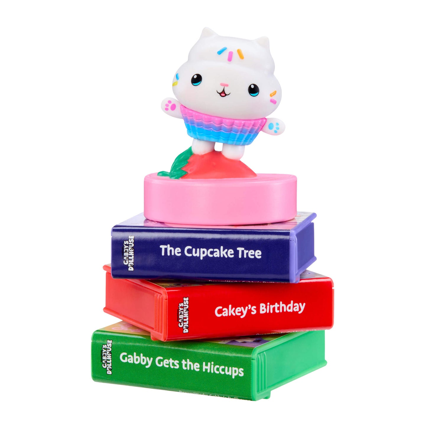 A children's book-themed toy stack featuring a white cat character wearing a pink and blue cupcake outfit, standing on top of four colorful books titled 'The Cupcake Tree', 'Cakey's Birthday', and 'Gabby Gets the Hiccups',