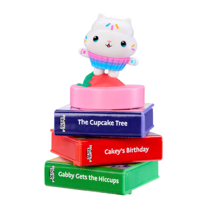 A children's book-themed toy stack featuring a white cat character wearing a pink and blue cupcake outfit, standing on top of four colorful books titled 'The Cupcake Tree', 'Cakey's Birthday', and 'Gabby Gets the Hiccups',