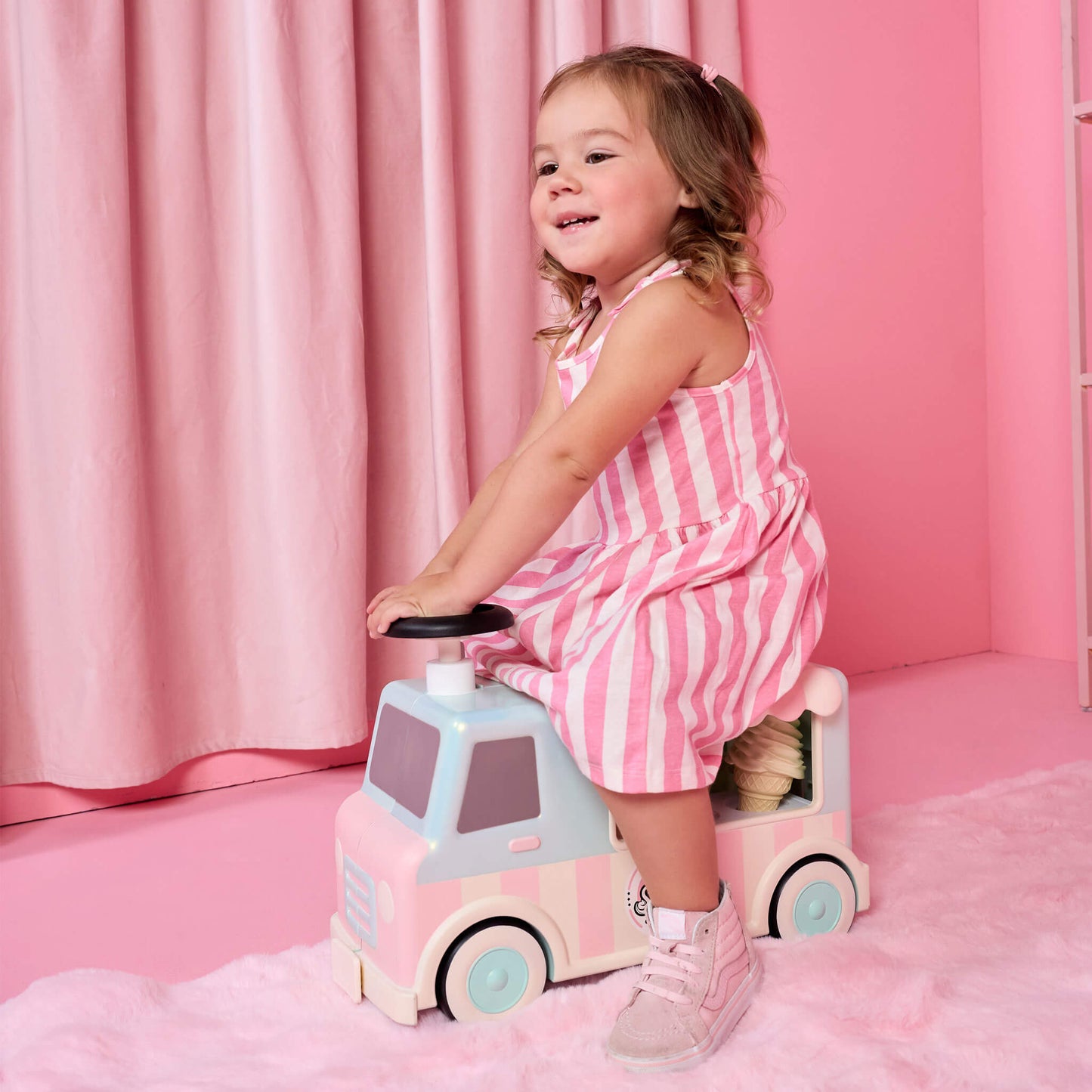 A young girl with blonde hair in a pink and white striped dress sits on a pastel pink and light blue toy truck in a pink room with a pink curtain backdrop.