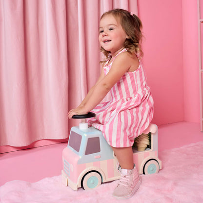 A young girl with blonde hair in a pink and white striped dress sits on a pastel pink and light blue toy truck in a pink room with a pink curtain backdrop.