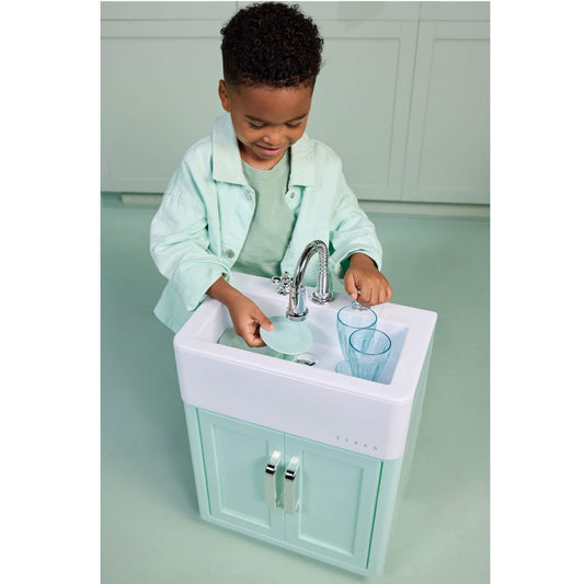 A young child wearing a mint green shirt and matching jacket plays with a mint green and white toy kitchen sink, washing dishes.