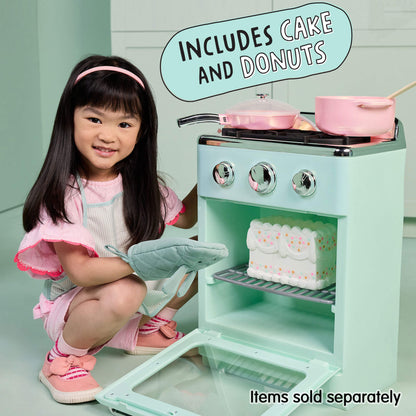 A young girl in a pink outfit kneels beside a mint green toy play stove, holding a gray oven mitt. The toy stove has silver knobs, pink pots, and a white cake on the shelf, with text overlays reading 'INCLUDES CAKE AND DONUTS' and 'Items sold separately'.