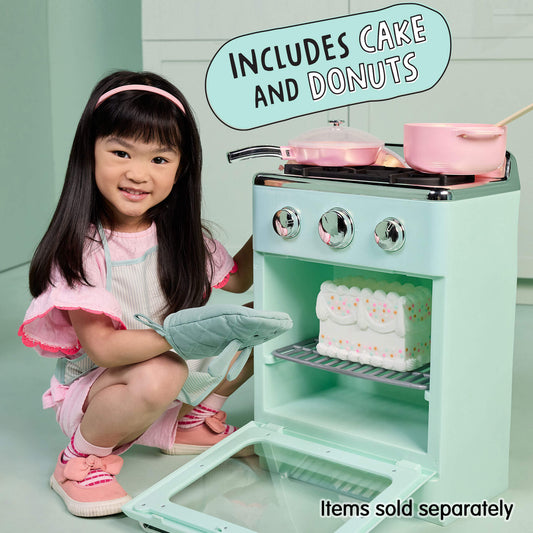 A young girl in a pink outfit kneels beside a mint green toy play stove, holding a gray oven mitt. The toy stove has silver knobs, pink pots, and a white cake on the shelf, with text overlays reading 'INCLUDES CAKE AND DONUTS' and 'Items sold separately'.