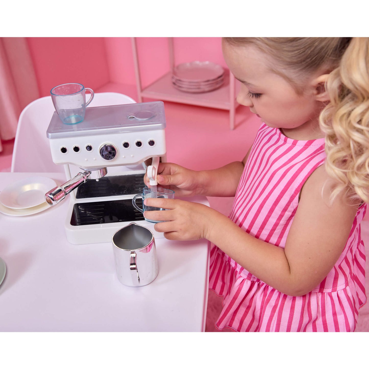 A young girl with blonde curly hair wearing a pink and white striped dress is playing with a white and silver toy espresso machine on a white table, making coffee with a plastic cup.