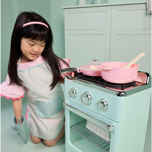 Girl with long dark hair wearing green apron with a pocket in front, kneeling by green stove with pink cook wear on top in a green kitchen setting