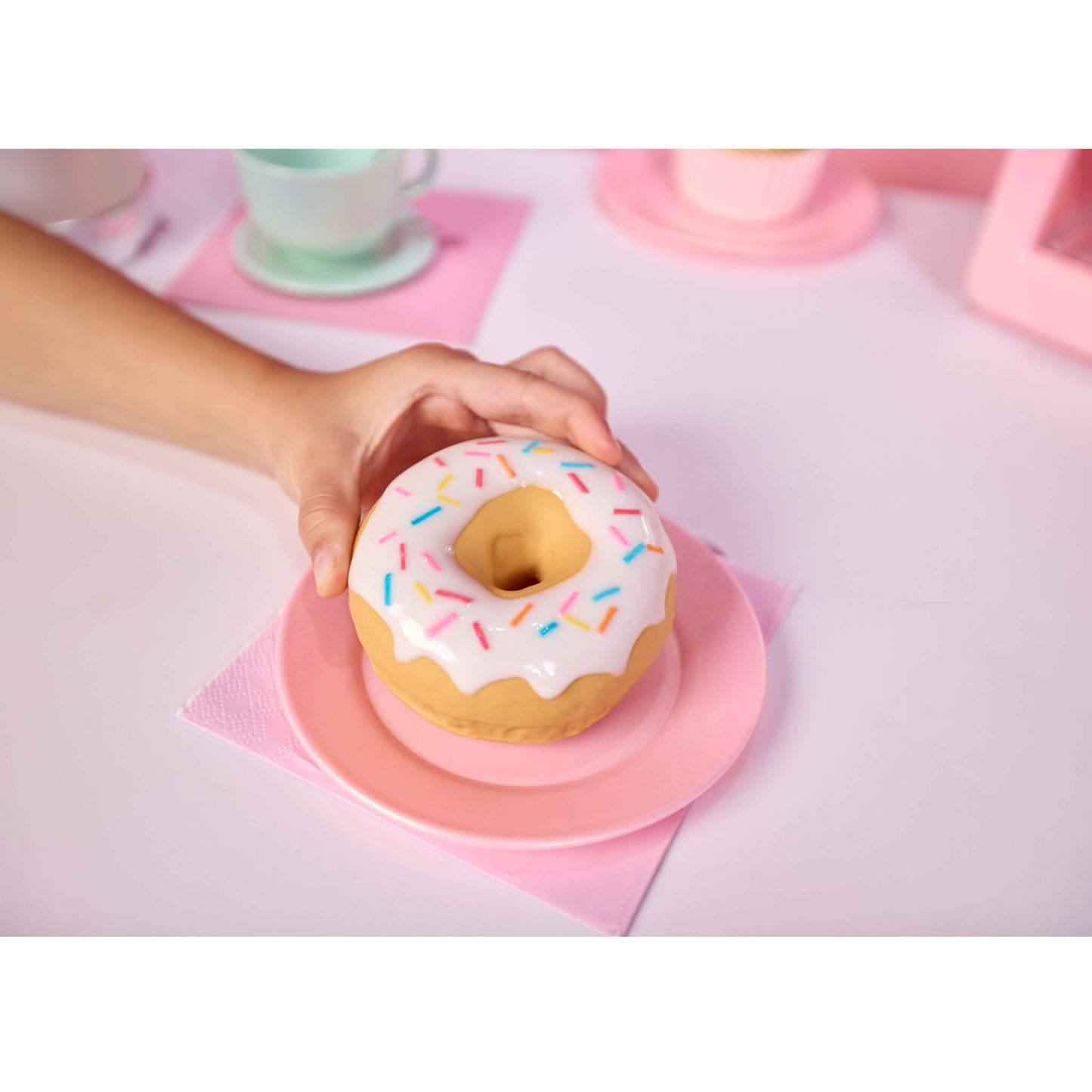 A hand holds a donut-shaped object with white frosting and multicolored sprinkles on a pink plate, set on a pastel pink table with cups in the background.