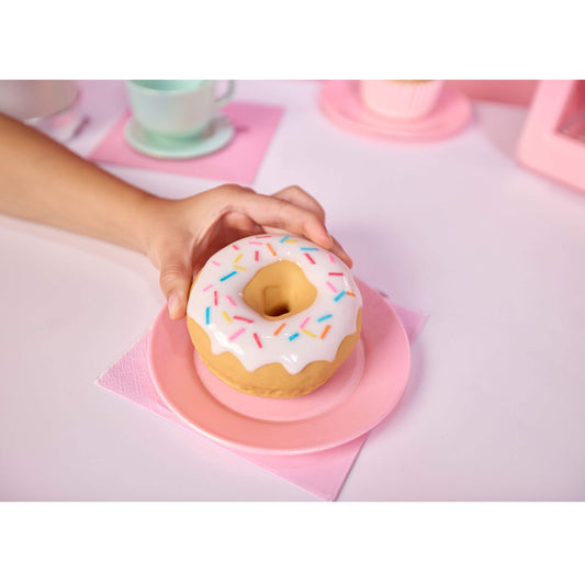 A hand holds a donut-shaped object with white frosting and multicolored sprinkles on a pink plate, set on a pastel pink table with cups in the background.