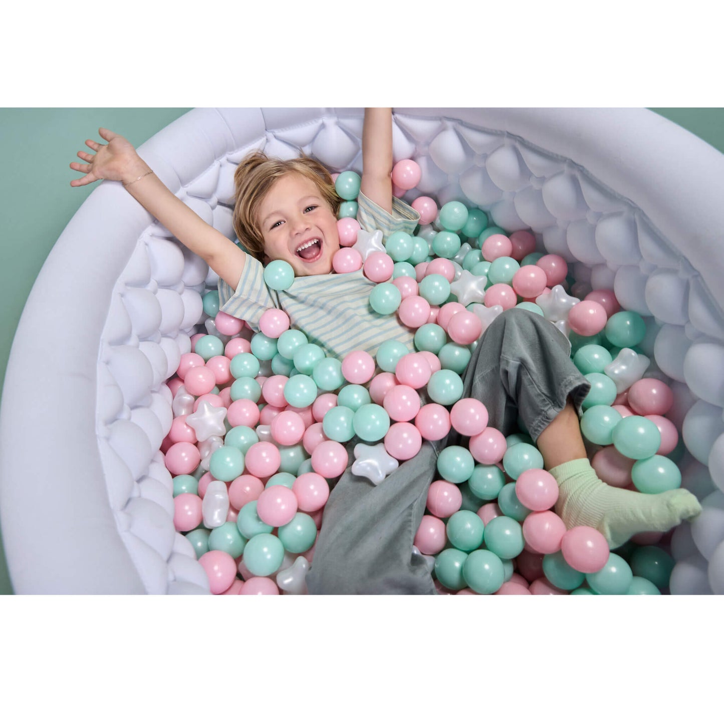 A young child with blonde hair lies happily inside a white ball pit filled with pastel pink and mint green plastic balls, wearing a striped shirt and gray pants.