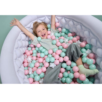 A young child with blonde hair lies happily inside a white ball pit filled with pastel pink and mint green plastic balls, wearing a striped shirt and gray pants.