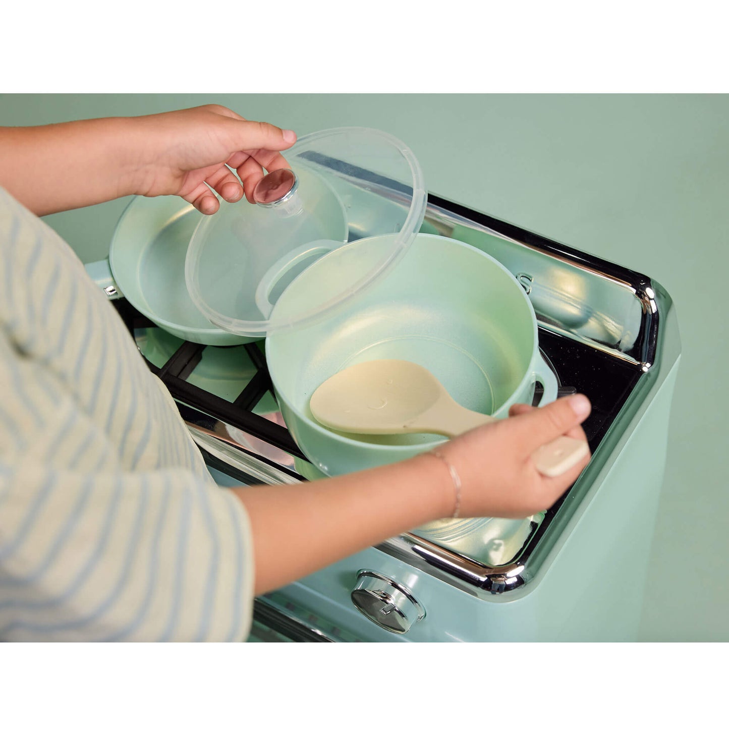 A child's hands are shown handling a mint green toy pot with a clear lid and a beige spoon on a retro-style mint green stove.