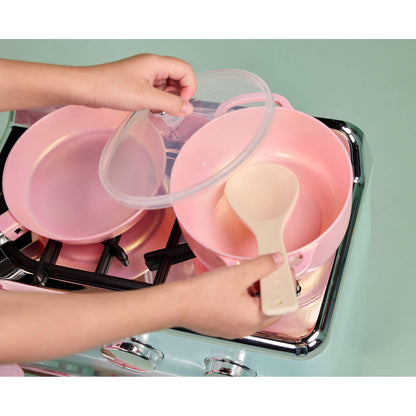 A pink cookware set on a toy stove, with a child's hands holding a clear lid and a beige spoon inside a pink pot.