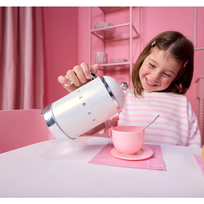 A young girl smiling while pouring water from a white and chrome kettle into a pink teacup and saucer on a table, set against a pink room background.