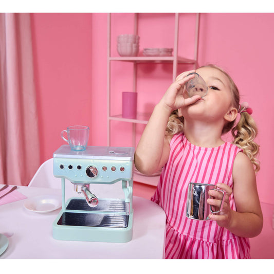 A young girl in a pink and white striped dress plays with a toy miniature Espresso machine and mugs on a white table against a pink background.