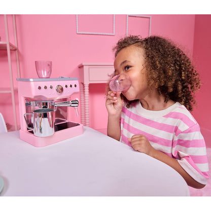 A young girl with curly brown hair wearing a pink and white striped t-shirt sits at a white table, drinking from a pink cup while a pink toy espresso machine with silver details sits on the table next to her.