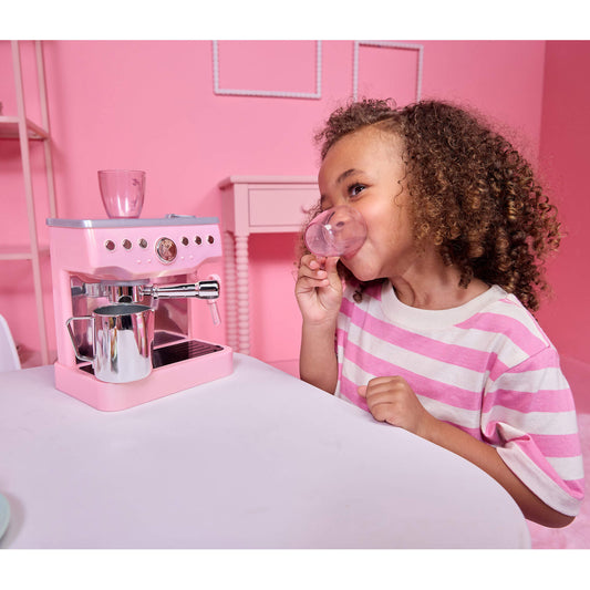 A young girl with curly brown hair wearing a pink and white striped t-shirt sits at a white table, drinking from a pink cup while a pink toy espresso machine with silver details sits on the table next to her.