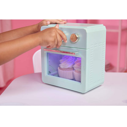 A child's hands are shown interacting with a toy retro-style mini oven toy. The toy oven is light mint green with a clear window showing purple light with cupcakes inside, set on a light pink table against a pink background.