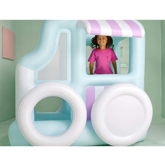 A child stands inside a large inflatable play structure shaped like a light blue and white truck in with pink-and-white striped awning details, photographed in a pastel-colored room.