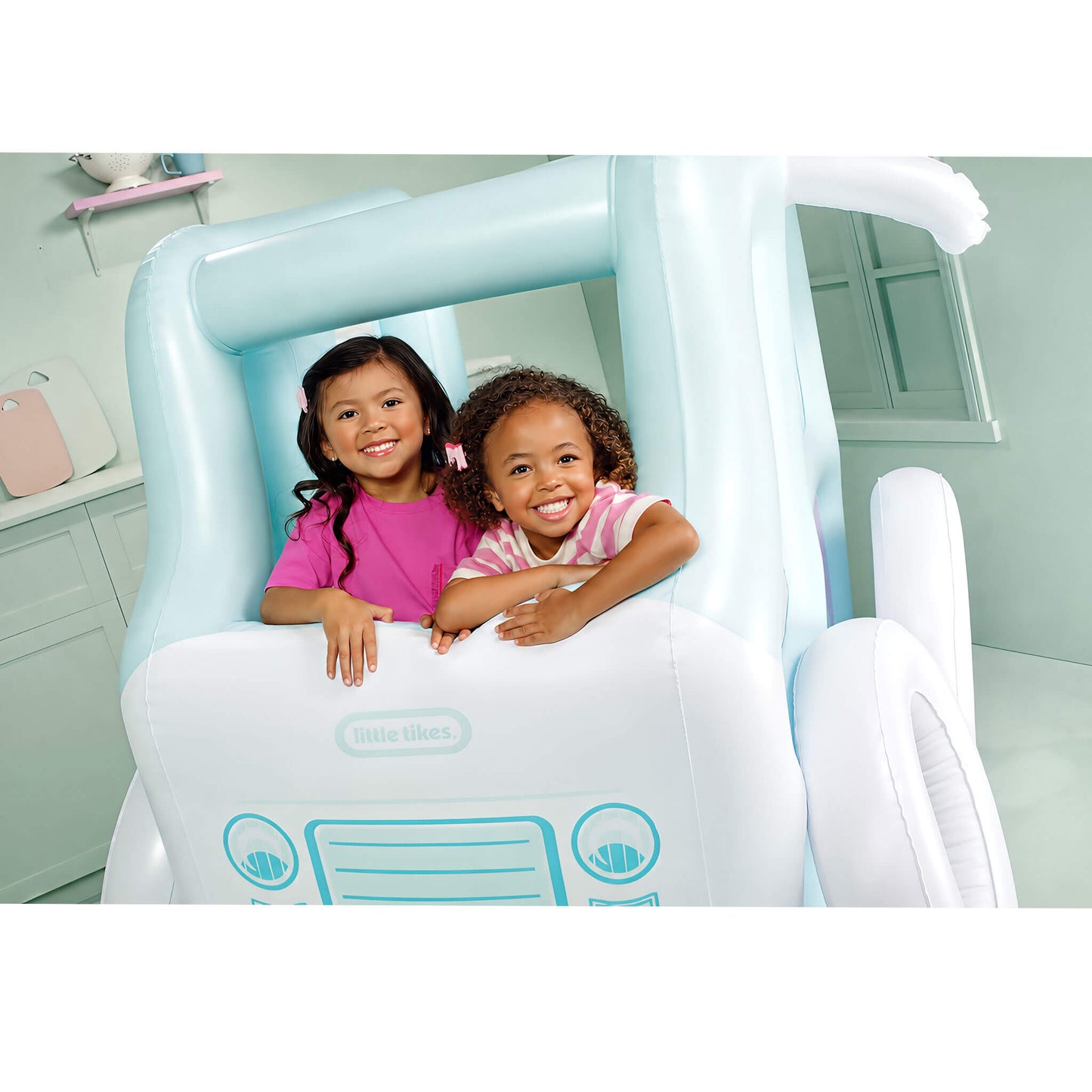 Two young girls smiling and posing inside a light blue and white inflatable truck-shaped, in a pale green room. The Little Likes brand logo is visible on the front of the inflatable.