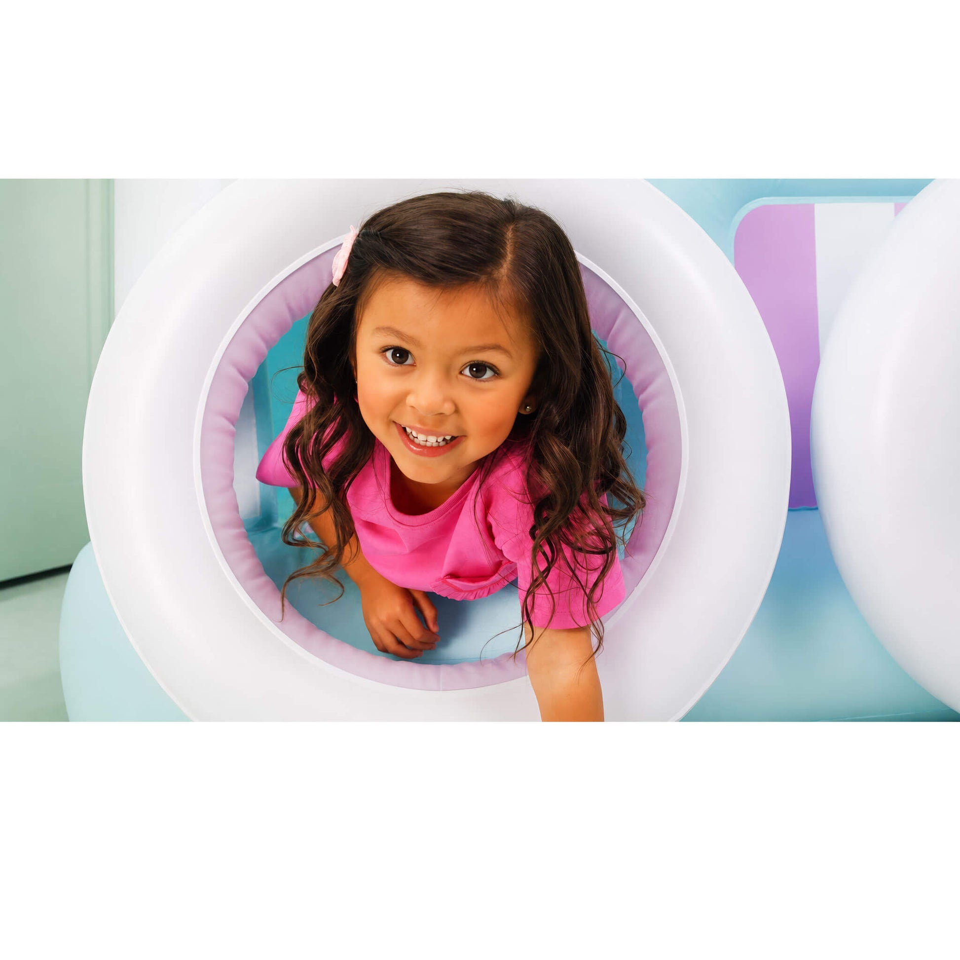 A young girl with long brown hair smiles while crawling inside a white inflatable tunnel with a pink padded interior, set against a light blue and purple play area.