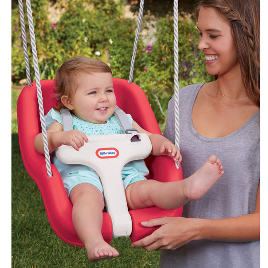 Baby in a red swing with a woman standing next to it outdoors.