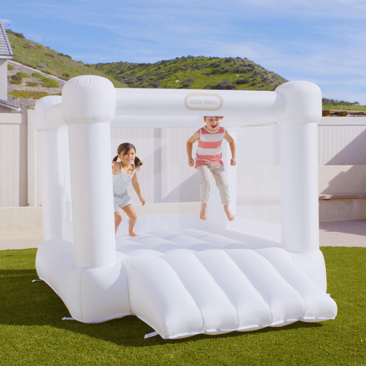 A children's inflatable bounce house set up on a green lawn outdoors, with two kids jumping inside. The product is white and a small visible Little Tikes logo on the top frame.