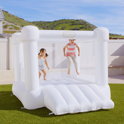 A children's inflatable bounce house set up on a green lawn outdoors, with two kids jumping inside. The product is white and a small visible Little Tikes logo on the top frame.
