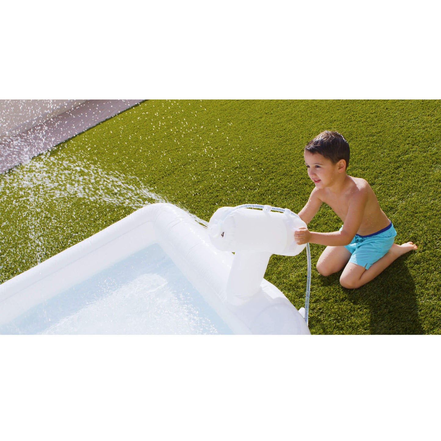 A young boy in blue swim trunks kneels beside a white inflatable pool sprayer, on a green grass lawn.